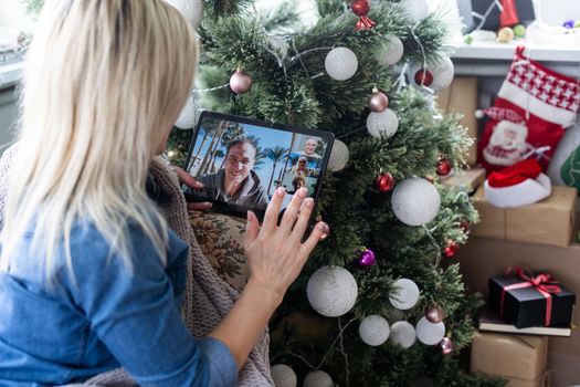 A young woman on the background of a Christmas tree with gifts, with a tablet has a video call or video chat with friends, family. Christmas online holiday. Stay home vacation.