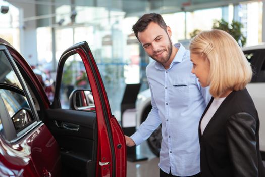 Handsome bearded man talking to his wife while choosing new car to buy at the dealership. Couple examining automobile on sale, copy space