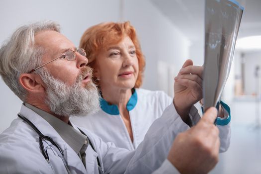 Close up of a bearded senior male doctor talking to his female co-worker, looking at MRI scan of a patient