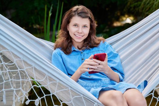 Outdoor portrait of teenage girl 12, 13 years old sitting in hammock with smartphone. Smiling girl looking at camera relaxing in backyard using mobile apps for study and leisure
