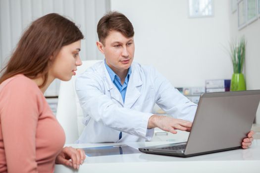 Mature male doctor showing something on laptop screen to his female patient during medical consultation