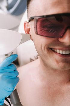 Cropped close up of a man smiling, getting laser hair removal treatment on his ear. Beautician removing hair on the ear of a male client, using laser