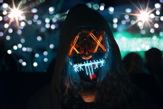 Person in hood with long disheveled hair in a lighting neon glowing mask. Anonymous man is a cyber criminal. Halloween dress-up party in night club. All Saints' Night.