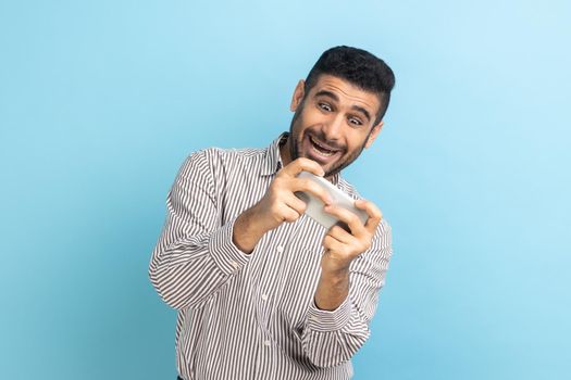 Delighted bearded businessman using mobile phone with happy positive expression, playing video game on cellphone, wearing striped shirt. Indoor studio shot isolated on blue background.