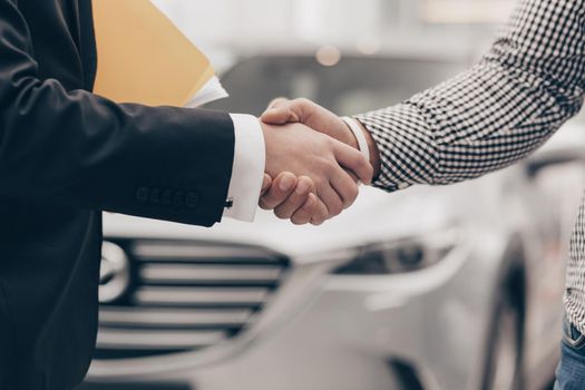 Close up cropped shot of a man shaking hands with professional car dealer at automobile dealership. Salesman shaking hands with his client after selling vehicle. Transportation, partnership concept