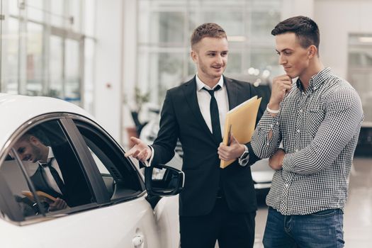 Professional car dealer showing cars on sale to his male customer, working at the auto dealership salon. Man looking thoughtfully at the car he is examining. Handsome man buying new automobile