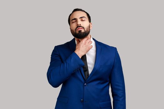 Unhealthy bearded man touching neck feeling pain while swallowing, result of chocking, throat inflammation, sore throat, wearing official style suit. Indoor studio shot isolated on gray background.
