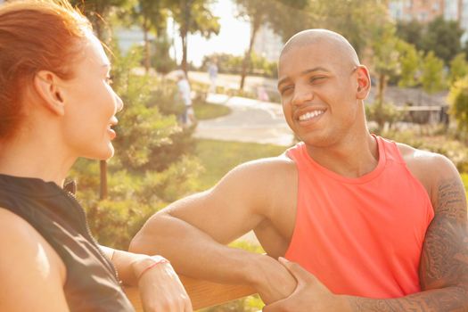 Happy handsome African man laughing, talking to his girlfriend while resting outdoors on a warm sunny day. Beautiful couple resting on the bench after exercising together