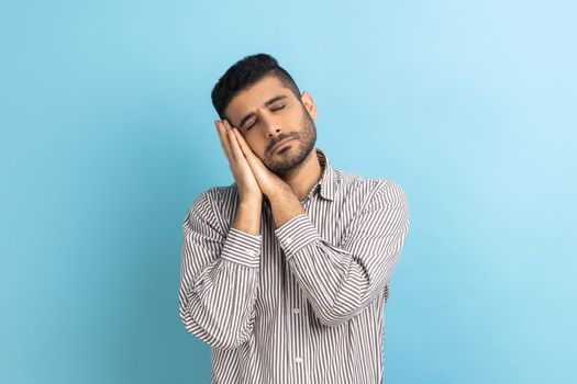 Portrait of handsome bearded man sleeping laying down on her palms, having comfortable nap and resting, dozing off, wearing striped shirt. Indoor studio shot isolated on blue background.