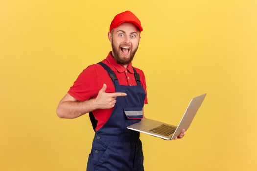 Side view of excited worker man wearing blue overalls, red T-shirt and cap working on laptop, pointing at screen with finger, looking at camera. Indoor studio shot isolated on yellow background.