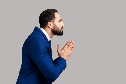 Side view of man holding hands in prayer, looking with imploring pleading expression, begging help, asking forgiveness, wearing official style suit. Indoor studio shot isolated on gray background.