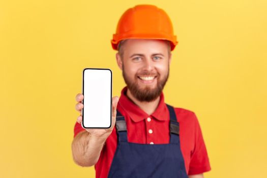 Portrait of happy smiling builder man wearing blue uniform and protective helmet holding smart phone with blank screen for advertisement. Indoor studio shot isolated on yellow background.