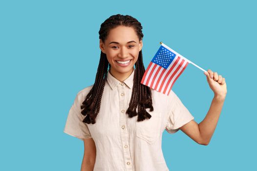 Smiling patriotic woman with dreadlocks holding in hand flag of united states of america, celebrating independence day, wearing white shirt. Indoor studio shot isolated on blue background.