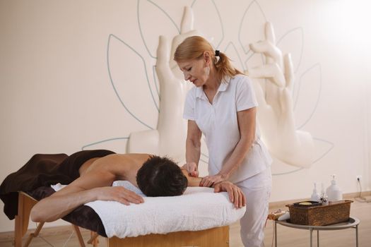 Senior female masseur working at spa center, massging male client, copy space