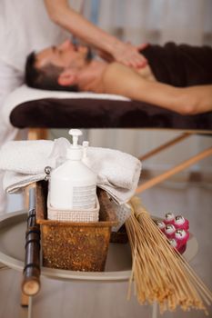 Vertical close up of skin moisturizer bottle and massage tools, man getting professional spa massage on background