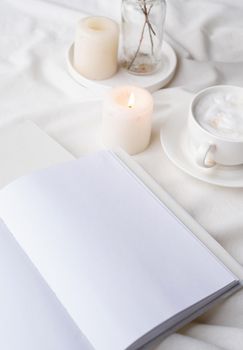 white blank book with cup of coffee and burning candle , cozy composition with opened book mockup on bed