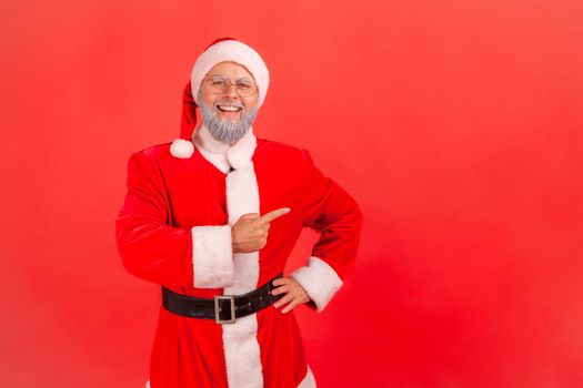 Satisfied elderly man with gray beard wearing santa claus costume standing with hand on hips, pointing index finger aside, presenting copy space. Indoor studio shot isolated on red background.