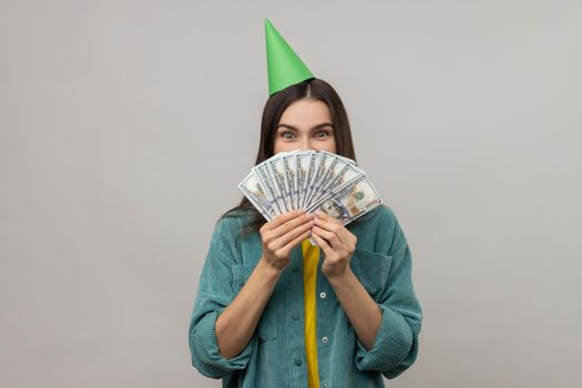 Portrait of shy woman with party cone on head covering half of face with money, lottery win, big profit, wearing casual style jacket. Indoor studio shot isolated on gray background.