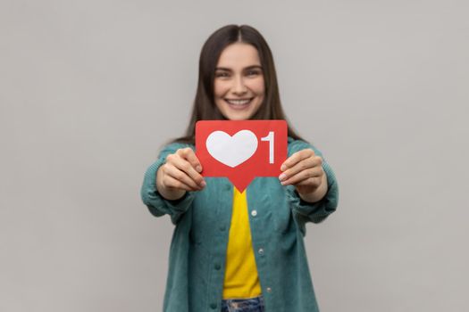 Satisfied woman holding out heart icon, recommending to follow and share blog with interesting content, wearing casual style jacket. Indoor studio shot isolated on gray background.