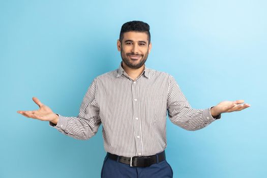Open hearted generous businessman with beard sharing opening hands looking at camera with kind smile, greeting and regaling, wearing striped shirt. Indoor studio shot isolated on blue background.