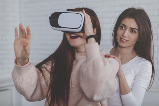 Charming young woman smiling, while her friend wearing 3d virtual reality headset, copy space. Excited woman trying virtual reality glasses for the first time. Female friends having fun, using vr goggles