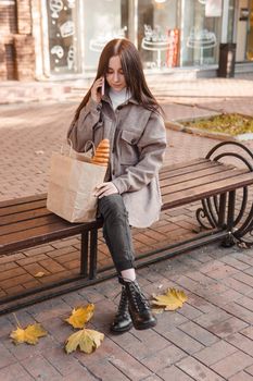 A stylish brunette woman walks around the autumn city. The brunette is sitting on a bench outside with a glass of coffee and a phone, holding a craft bag with a baguette.