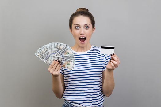 Portrait of shocked woman wearing striped T-shirt holding credit card and dollar banknotes, looking at camera with big eyes, surprised with easy banking. Indoor studio shot isolated on gray background
