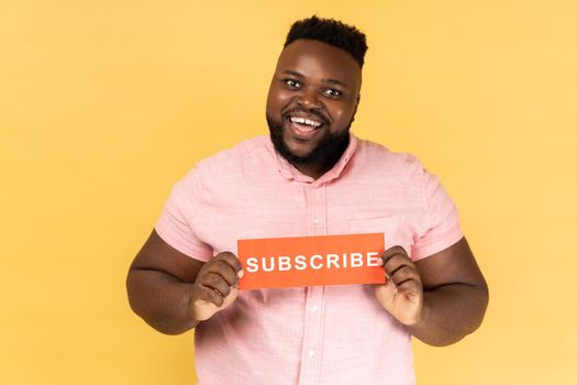 Portrait of cheerful positive bearded man wearing pink shirt holding red card with subscribe inscription, asking to follow his vlog. Indoor studio shot isolated on yellow background.