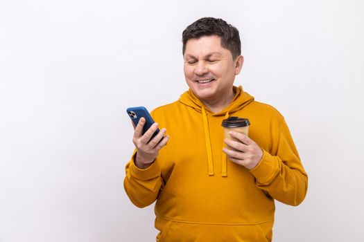 Portrait of satisfied handsome man standing with coffee and chatting on the phone, expressing positive emotions, wearing urban style hoodie. Indoor studio shot isolated on white background.