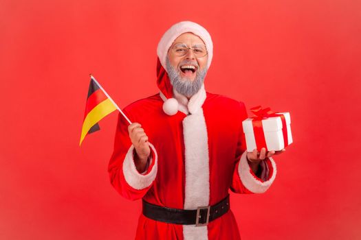 Portrait of happy excited elderly man with gray beard wearing santa claus costume holding German flag and box with Christmas present. Indoor studio shot isolated on red background.