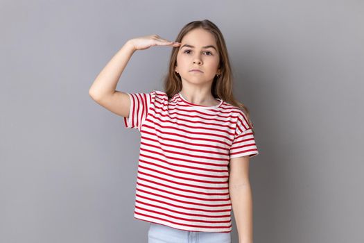 Portrait of serious bossy dark haired little girl wearing striped T-shirt standing, making salute with hand on head, accepting order. Indoor studio shot isolated on gray background.