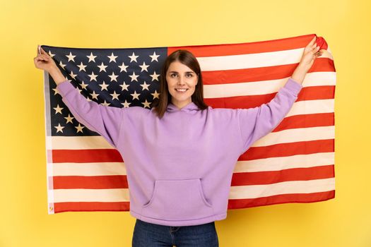 Portrait of attractive happy positive woman standing with raised arms, holding USA flag, celebrating national holiday, wearing purple hoodie. Indoor studio shot isolated on yellow background.