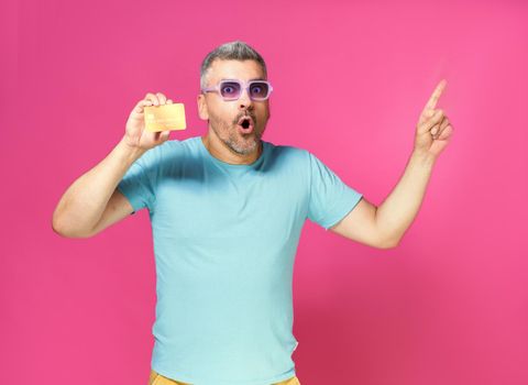 Look at this great offer, grey haired man hold debit, credit card in hand pointing sideways wearing blue t-shirt and purple glasses isolated on pink background. Financial, banking concept.