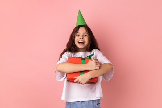 Portrait of excited little girl wearing white T-shirt and party cone embracing red gift box, getting present from friend on her birthday. Indoor studio shot isolated on pink background.