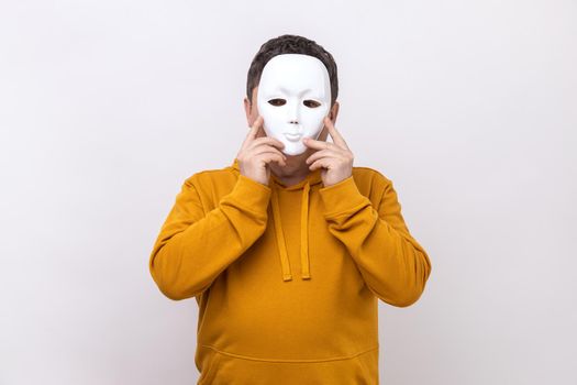 Portrait of unknown middle aged man covering her face with white mask, hiding her real personality, anonymity, wearing urban style hoodie. Indoor studio shot isolated on white background.