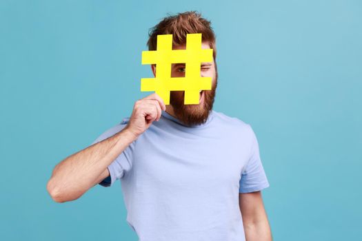 Portrait of bearded man hiding face behind yellow hashtag sign, calls on to tag topics on websites, giving recommendations, peeping. Indoor studio shot isolated on blue background.