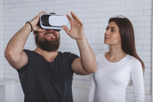 Bearded man using virtual reality headset, his girlfriend watching him. Young couple trying 3d vr glasses at work together. Friends using virtual reality goggles, copy space. Futuristic concept