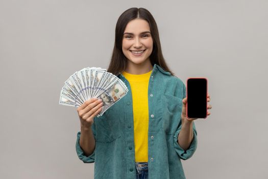 Smiling woman holding in hands smart phone with blank screen and dollars banknotes, copy space for advertisement, wearing casual style jacket. Indoor studio shot isolated on gray background.