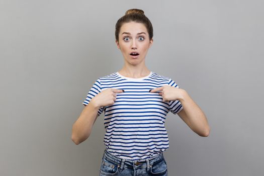 Portrait of astonished woman wearing striped T-shirt pointing at herself, asks who me, has surprised expression, shocked being picked. Indoor studio shot isolated on gray background.