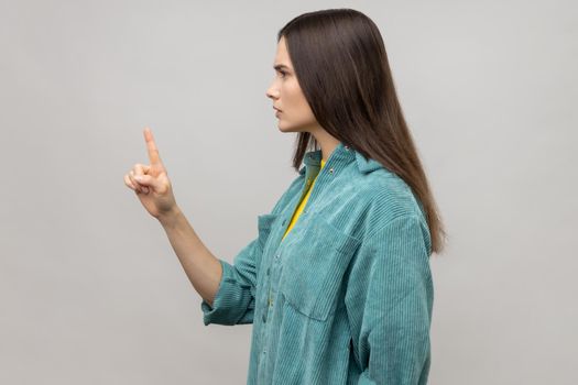 Be careful. Side view of strict bossy woman looking at camera seriously, pointing finger and warning, wearing casual style jacket. Indoor studio shot isolated on gray background.