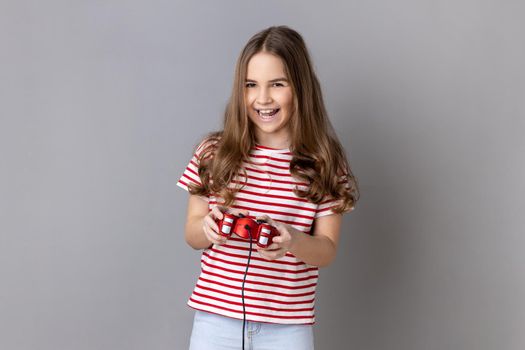 Portrait of little girl wearing striped T-shirt holding in hands red gamepad joystick, looking at camera with optimistic expression, enjoying game. Indoor studio shot isolated on gray background.