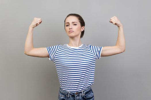 Portrait of proud woman wearing striped T-shirt showing biceps, expressing power ambitions to become successful, female rights, feminism. Indoor studio shot isolated on gray background.
