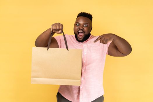 Portrait of excited bearded man wearing pink shirt pointing at shopping bag, looking at camera with pleased happy facial expression. Indoor studio shot isolated on yellow background.