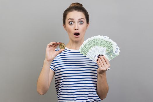 Portrait of surprised amazed shocked woman wearing striped T-shirt holding golden coin of crypto currency and big fan of euro banknotes. Indoor studio shot isolated on gray background.