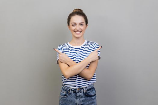 Portrait of satisfied woman wearing striped T-shirt pointing both sides, showing blank copy space for idea presentation, commercial text, toothy smile. Indoor studio shot isolated on gray background.