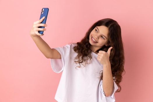 Portrait of satisfied little girl wearing white T-shirt showing thumbs up and smiling looking at camera of his smartphone, blogging. Indoor studio shot isolated on pink background.