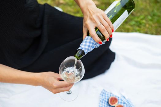 Romantic picnic in the park on the grass, delicious food: basket, wine, grapes, figs, cheese, blue checkered tablecloth, two glasses of wine. Girl pours wine into a glass.Outdoor recreation concept