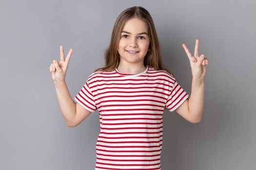 Portrait of positive optimistic adorable little girl wearing striped T-shirt gesturing victory sign at camera, success or achievement. Indoor studio shot isolated on gray background.