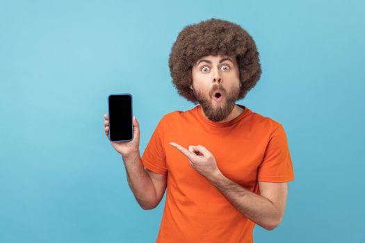 Portrait of shocked satisfied man with Afro hairstyle wearing orange T-shirt pointing at mobile phone with finger, showing empty space for advertisement. Indoor studio shot isolated on blue background