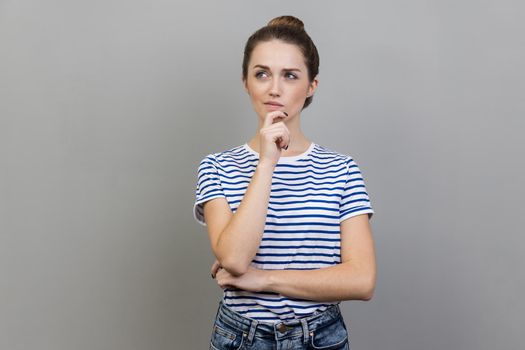 Portrait of pensive woman wearing striped T-shirt standing holding chin, thinking about important question, having thoughtful facial expression. Indoor studio shot isolated on gray background.